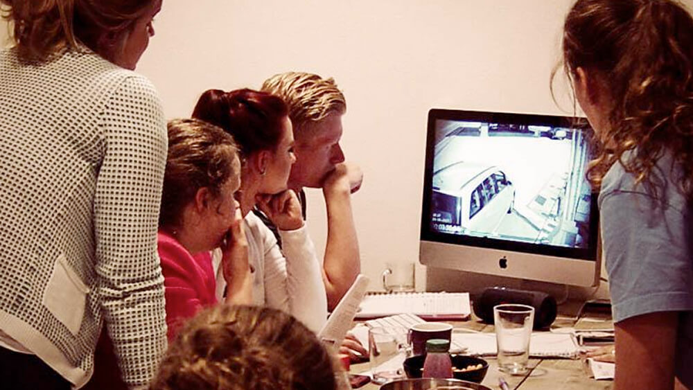 foto van mensen die aan een tafel naar een computerscherm kijken waar bewakingsbeelden op getoond worden voor een moordspel thuis